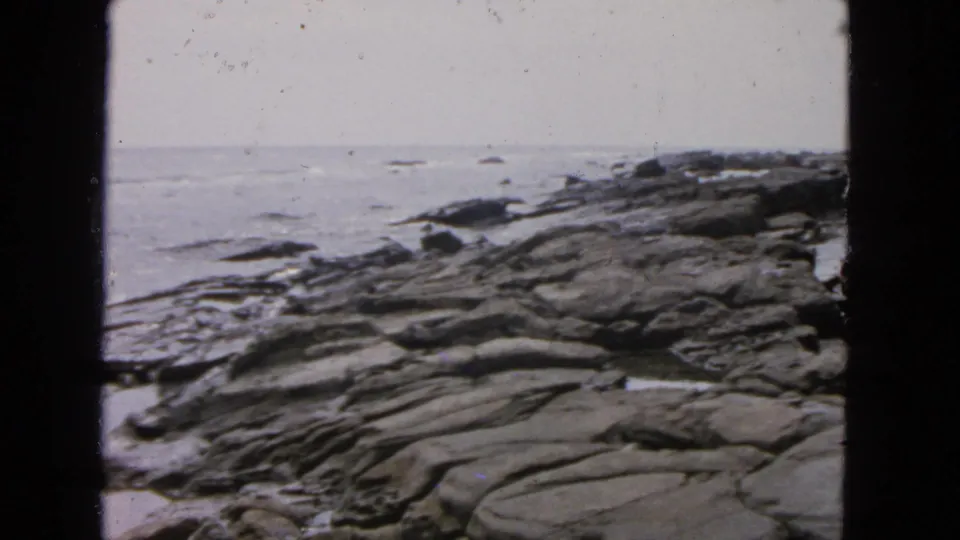 1963 San Francisco coastal home-movie frame with dark shoreline rocks and gentle sea beyond.