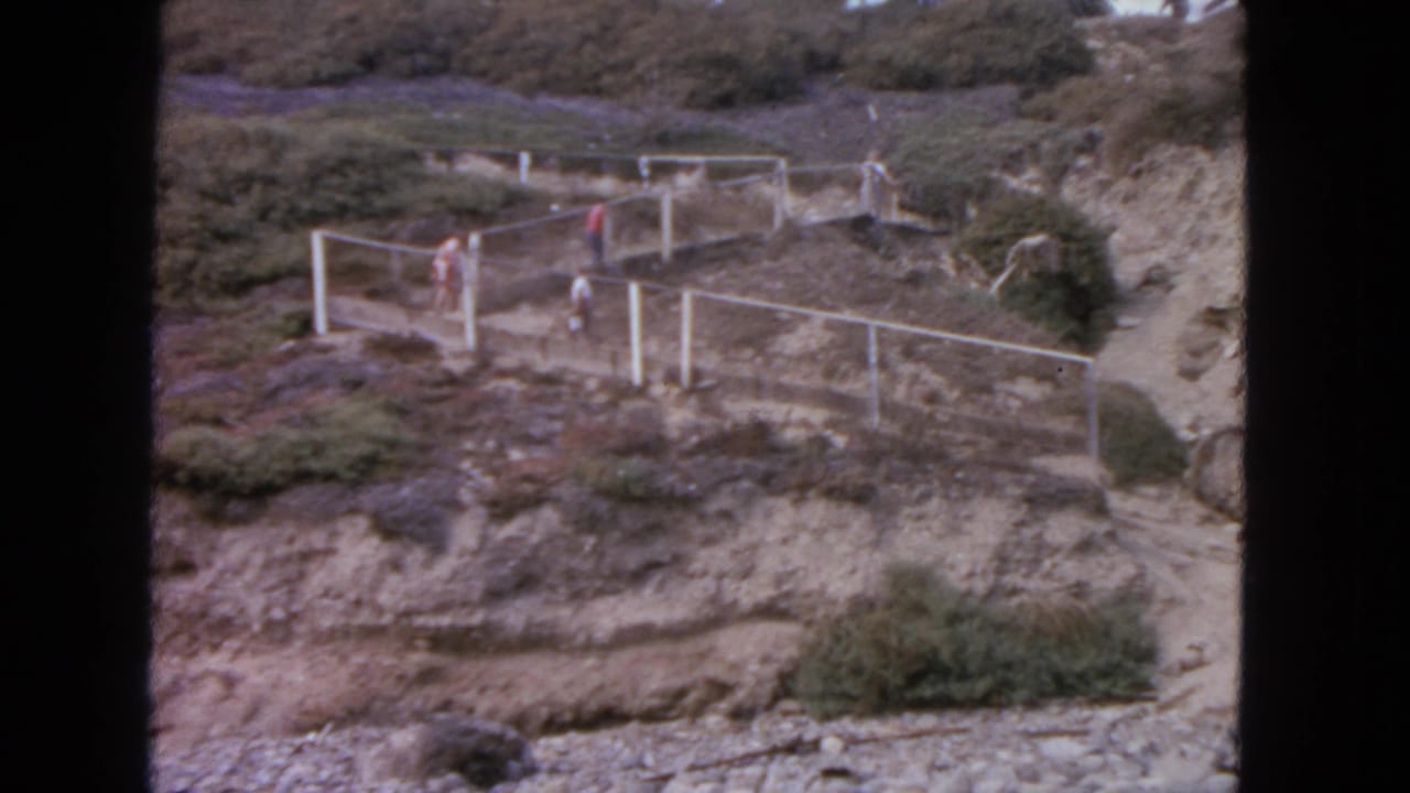 1963 San Francisco home-movie frame showing people moving along a fenced hillside path in a rugged, shrub-covered landscape.