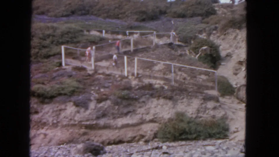 1963 San Francisco home-movie frame showing people moving along a fenced hillside path in a rugged, shrub-covered landscape.