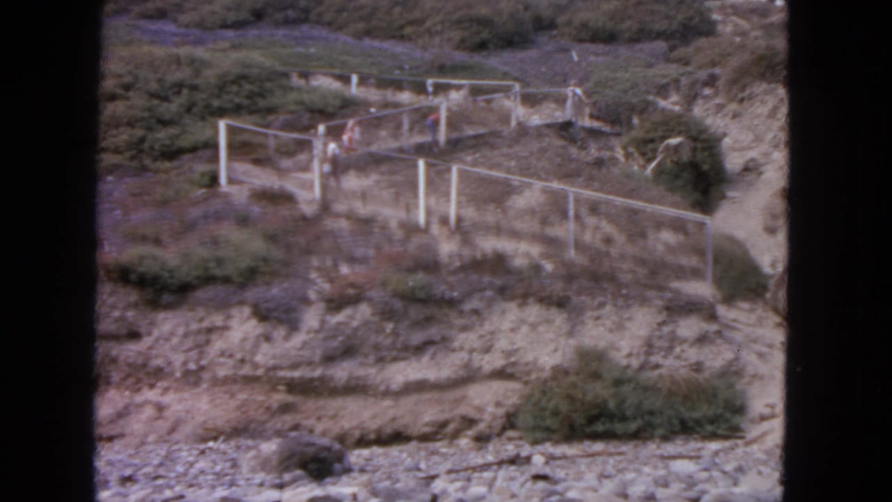 Distant people in a 1963 San Francisco outdoor scene walk beside pale fence rails on a rugged hillside path.