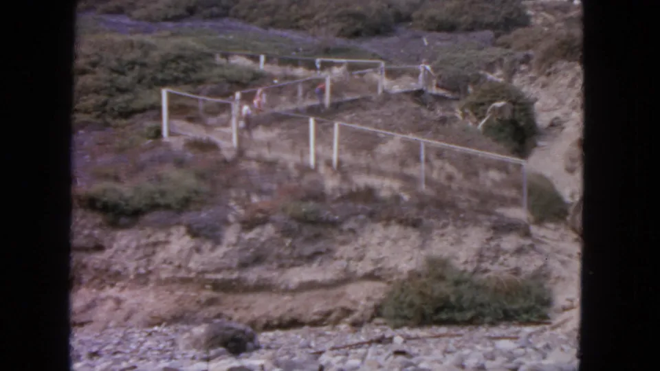 Distant people in a 1963 San Francisco outdoor scene walk beside pale fence rails on a rugged hillside path.