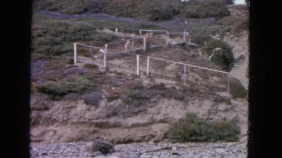 Home-movie view of several people crossing a fenced hillside route in San Francisco, with shrubs and exposed earth visible.