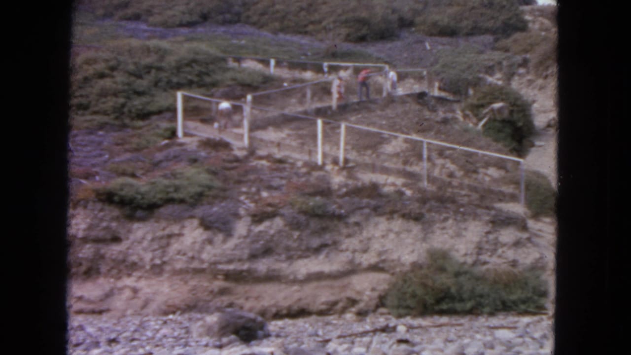 Distant people walk near a fenced path on a scrubby rocky hillside.