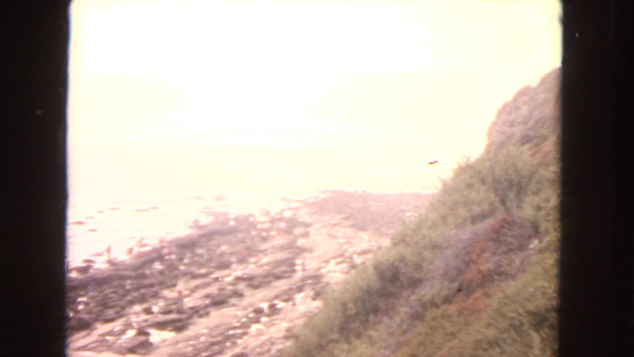 1963 San Francisco-area coastal view with a bright ocean horizon, rocky shore below, and a grassy hillside on the right.