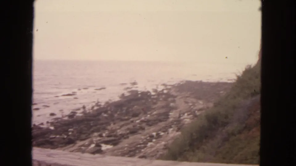 1963 8mm view from a coastal hillside, looking across a curving road toward rocks, tide pools, and the ocean.