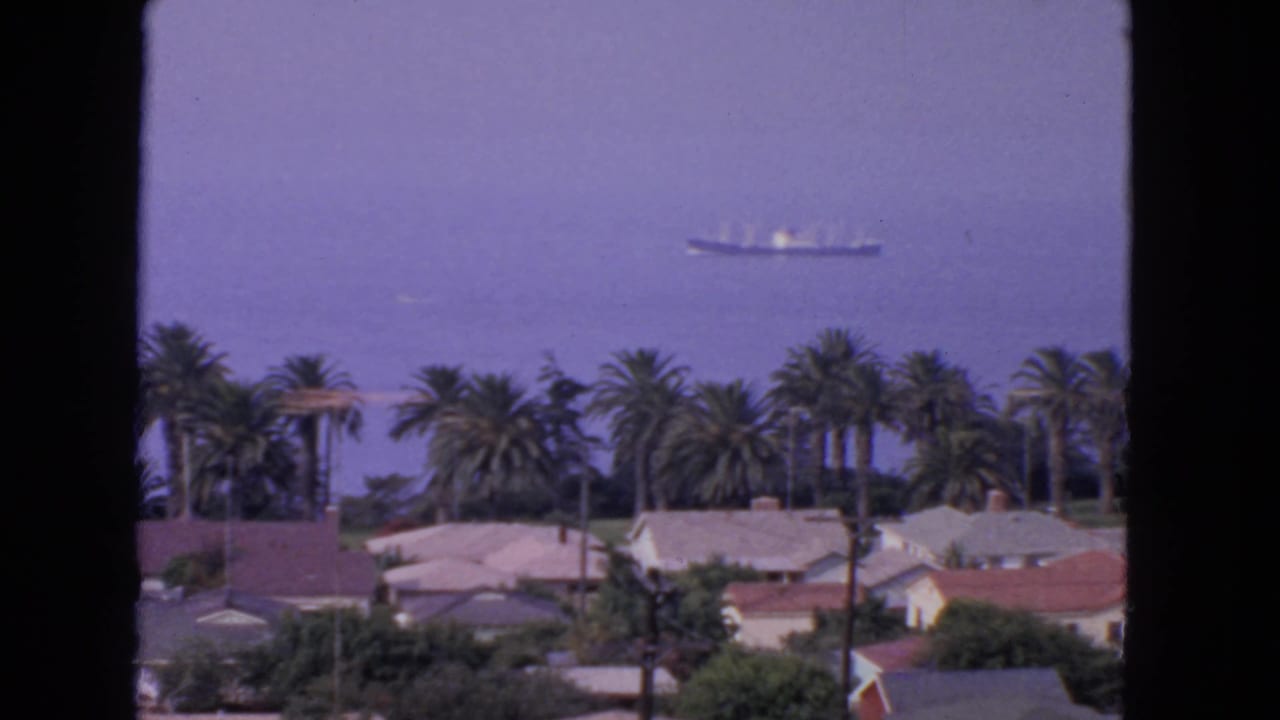 Cargo ship on hazy water above palm trees and rooftops, with dark frame borders.