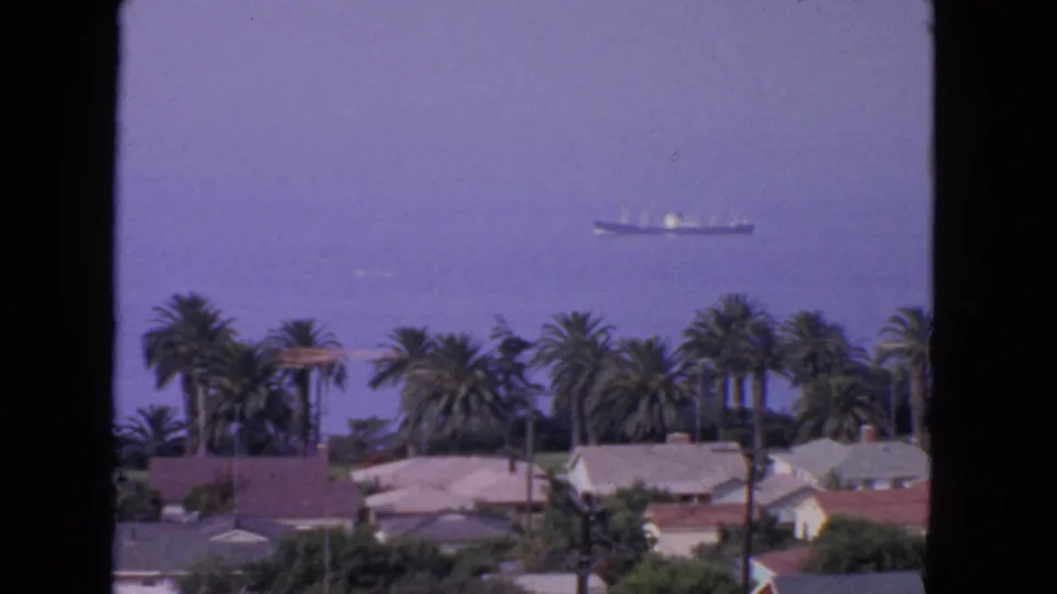 1963 San Francisco bay view with rooftops and palms below a distant cargo ship and small aircraft.