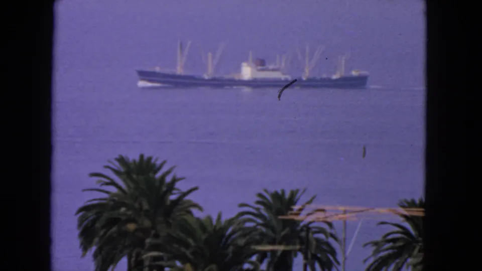Closer 1963 San Francisco home-movie frame of a cargo ship on the bay, with palms and rooftops in the foreground.