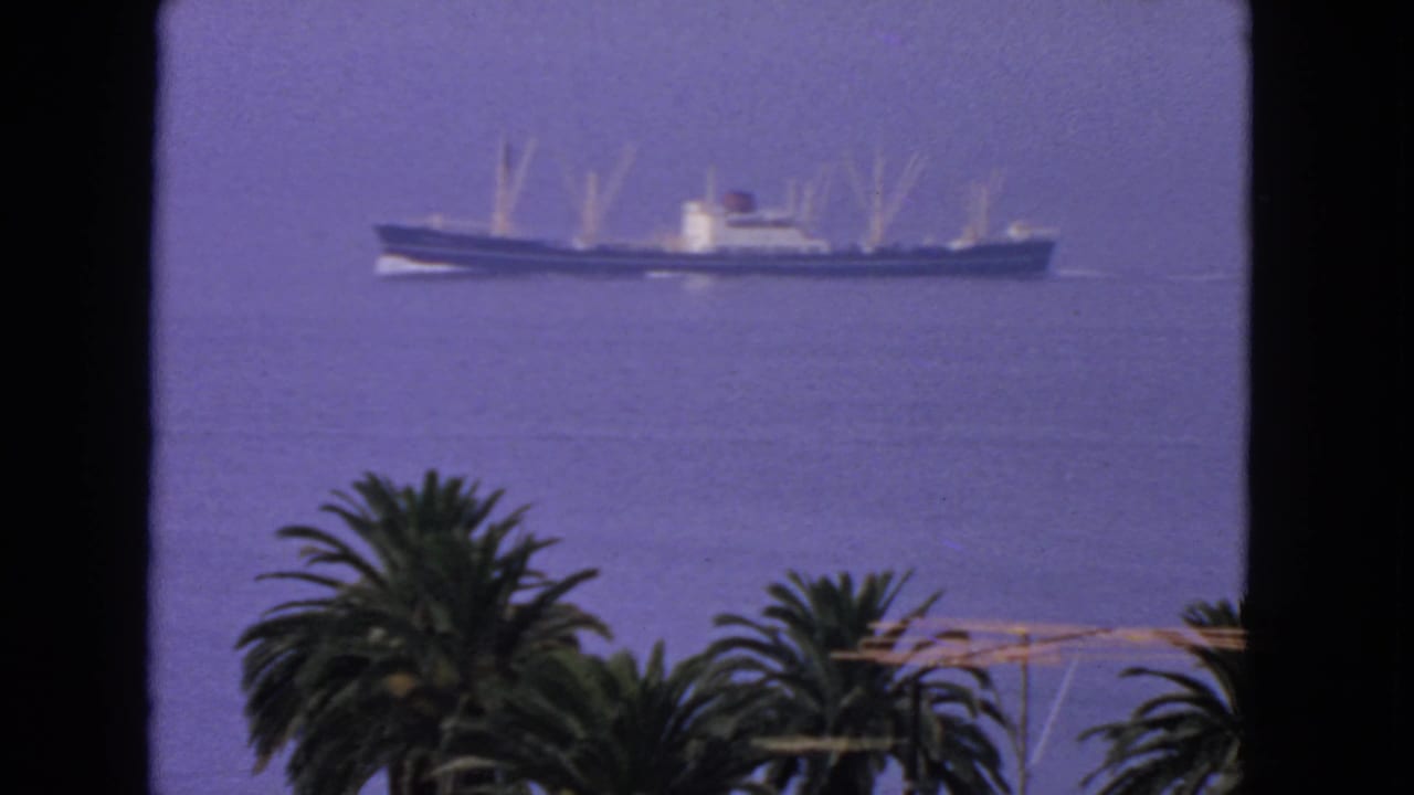 1963 San Francisco Bay home-movie frame showing a long ship on calm blue water beyond dark window edges and foreground palms.