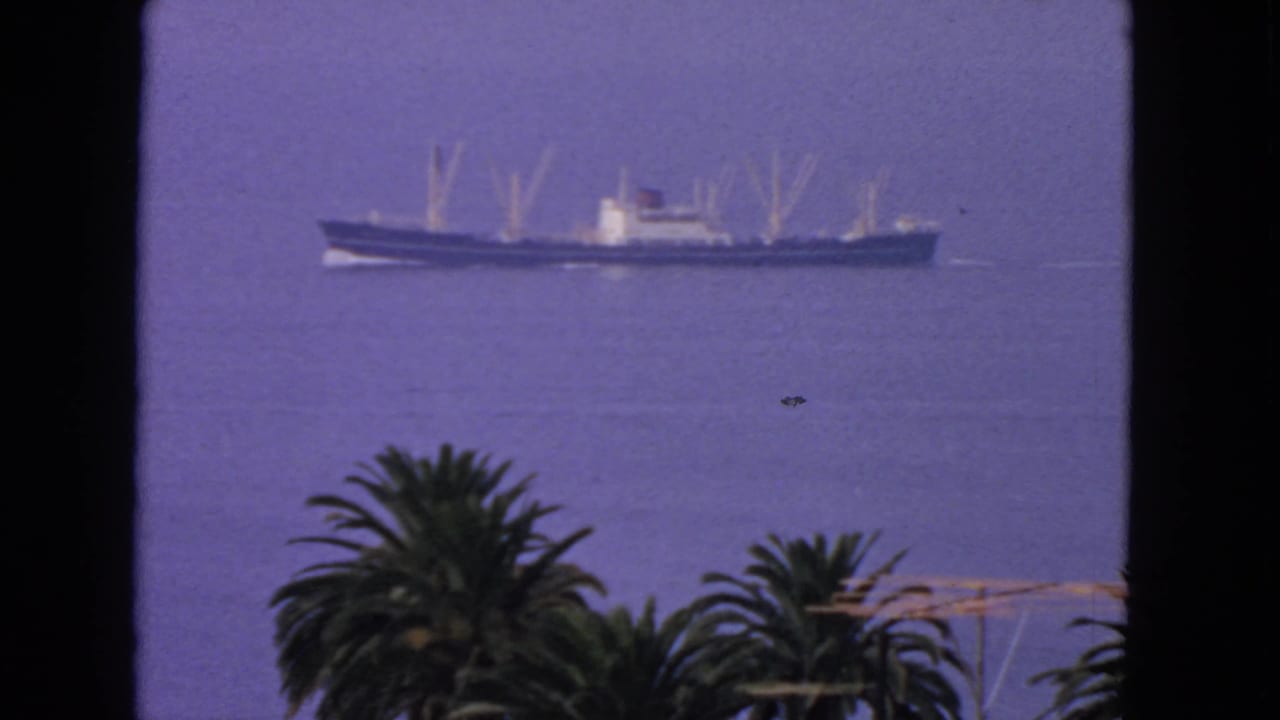 A long narrow ship moves across smooth San Francisco Bay water in a 1963 8mm frame, with palms and dark window edges in front.