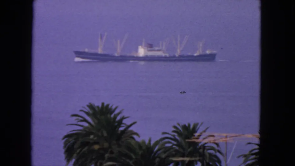 A long narrow ship moves across smooth San Francisco Bay water in a 1963 8mm frame, with palms and dark window edges in front.