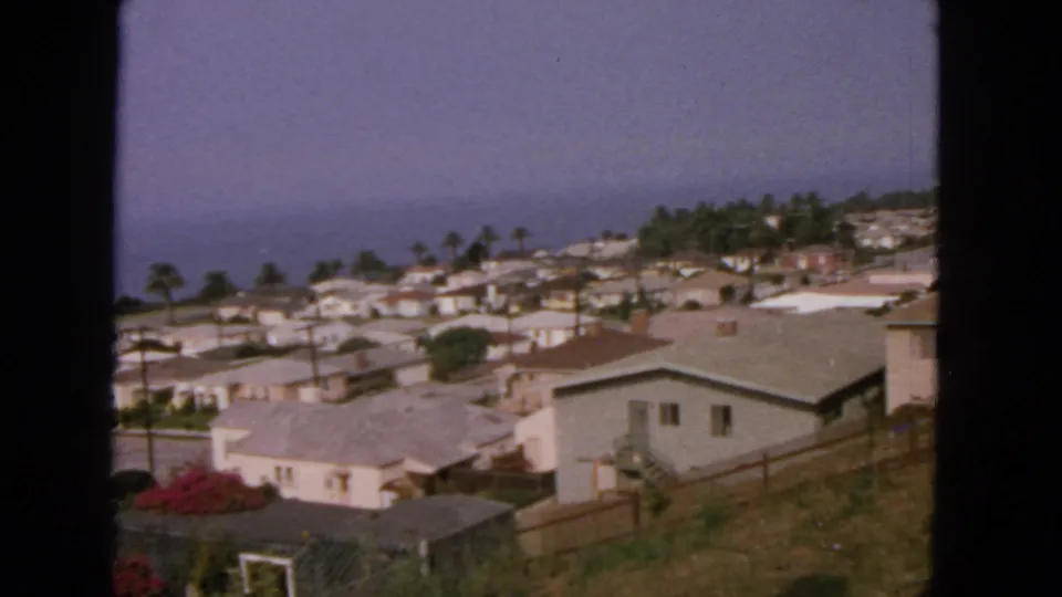 1963 San Francisco-area hillside neighborhood with mid-century homes, fences, palms, and a hazy coastal horizon beyond.