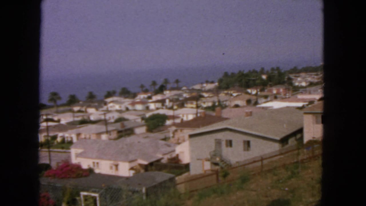 1963 home-movie view over a California coastal hillside neighborhood, with low houses, palms, and the ocean horizon beyond.