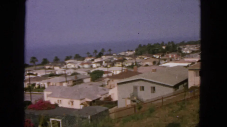 1963 home-movie view over a California coastal hillside neighborhood, with low houses, palms, and the ocean horizon beyond.