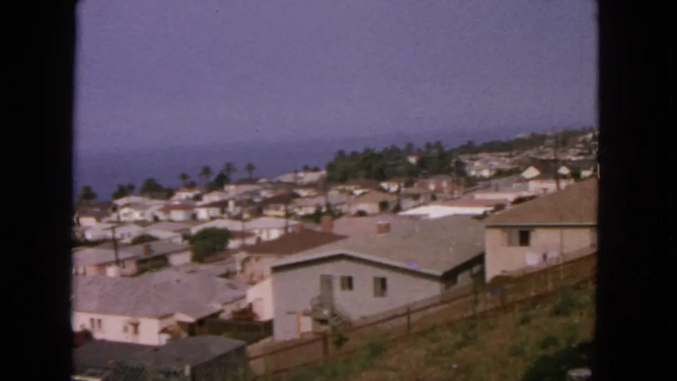 Home-movie frame showing a San Francisco-area coastal hillside neighborhood in 1963, with pale houses and palms below the ocean horizon.
