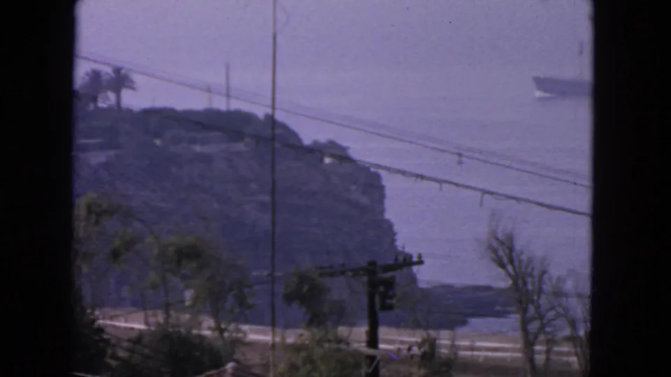 1963 coastal view near San Francisco showing a rocky bluff, utility lines, roadway, and a distant ship on the water.