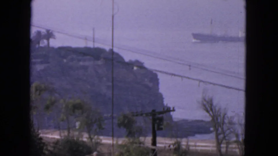 1963 San Francisco 8mm frame with a freighter-like vessel beyond a rocky coast, foreground wires, trees, and road infrastructure.