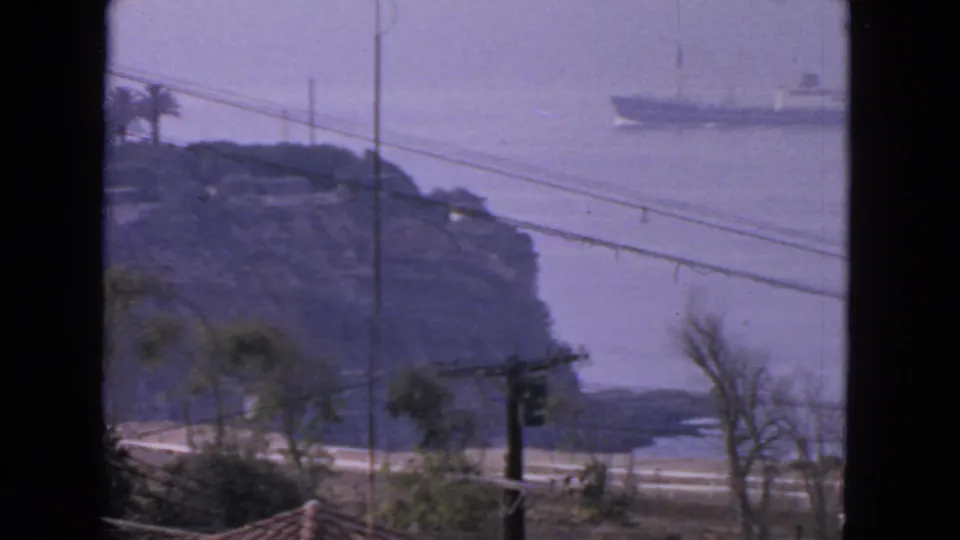 1963 San Francisco coastal home-movie still showing a distant ship, rocky bluff, utility lines, sparse trees, and hazy bay water.