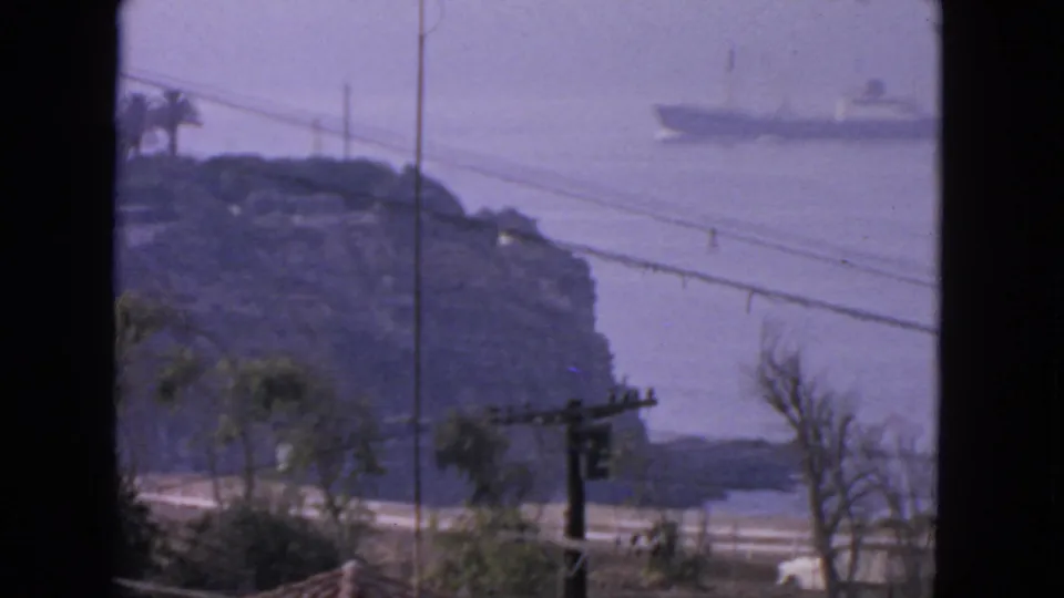 1963 San Francisco coastal home-movie frame showing a ship offshore beyond a rocky shoreline, road, utility lines, and sparse trees.
