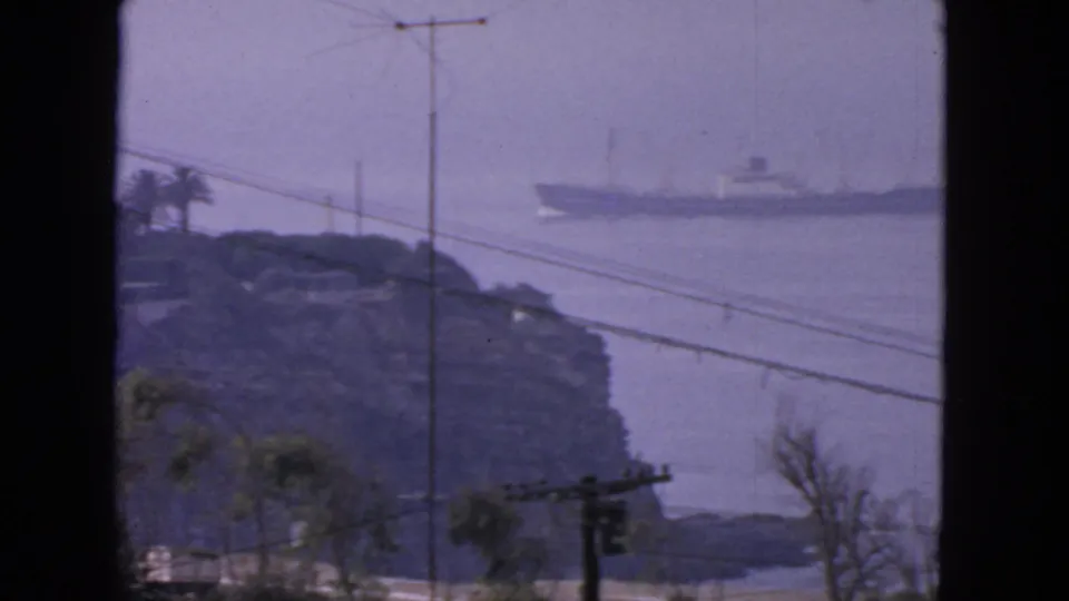1963 San Francisco coastal home-movie frame with a ship offshore, rocky shoreline, utility poles, crossing wires, and roadside trees.