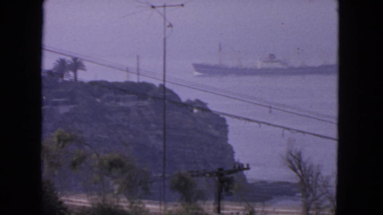 Distant ocean liner passes behind a rocky island with power lines and an antenna in the foreground.