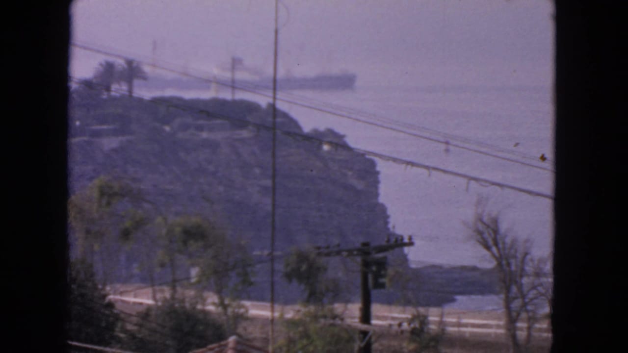 1963 San Francisco 8mm frame showing a hazy coastal bluff, distant merchant ship offshore, and foreground utility lines.
