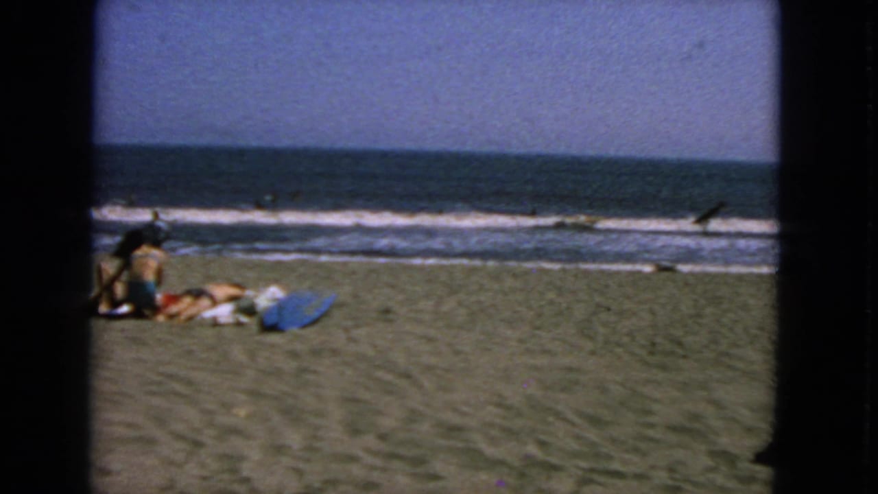 Beachgoers sit on sand near a blue towel or board with ocean waves behind them.