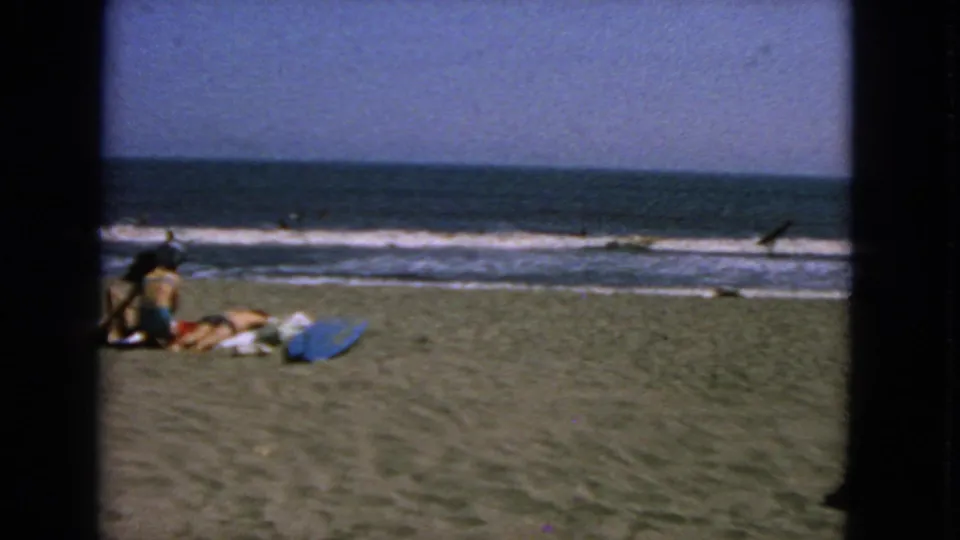 1963 San Francisco beach scene with people lounging on sand near a blue towel or board as waves roll in behind them.