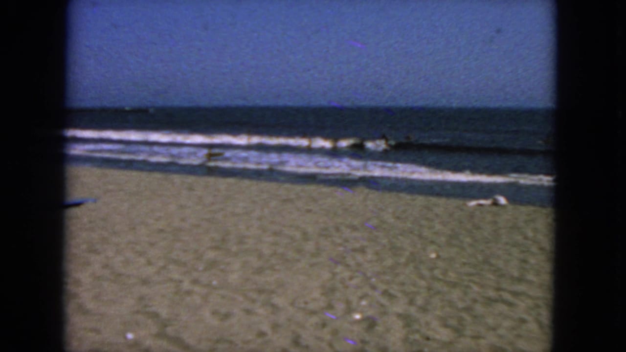 Wide 1963 San Francisco shoreline view with open sand, breaking surf, and small distant figures in the ocean.