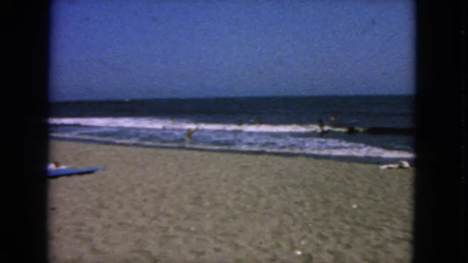 1963 San Francisco beach footage showing waves, scattered distant beachgoers, and a blue board or towel at the edge of frame.