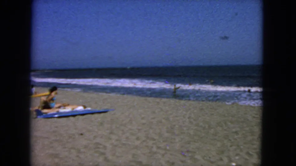 1963 San Francisco beach scene with two beachgoers seated on a blue towel or board as waves break behind them.