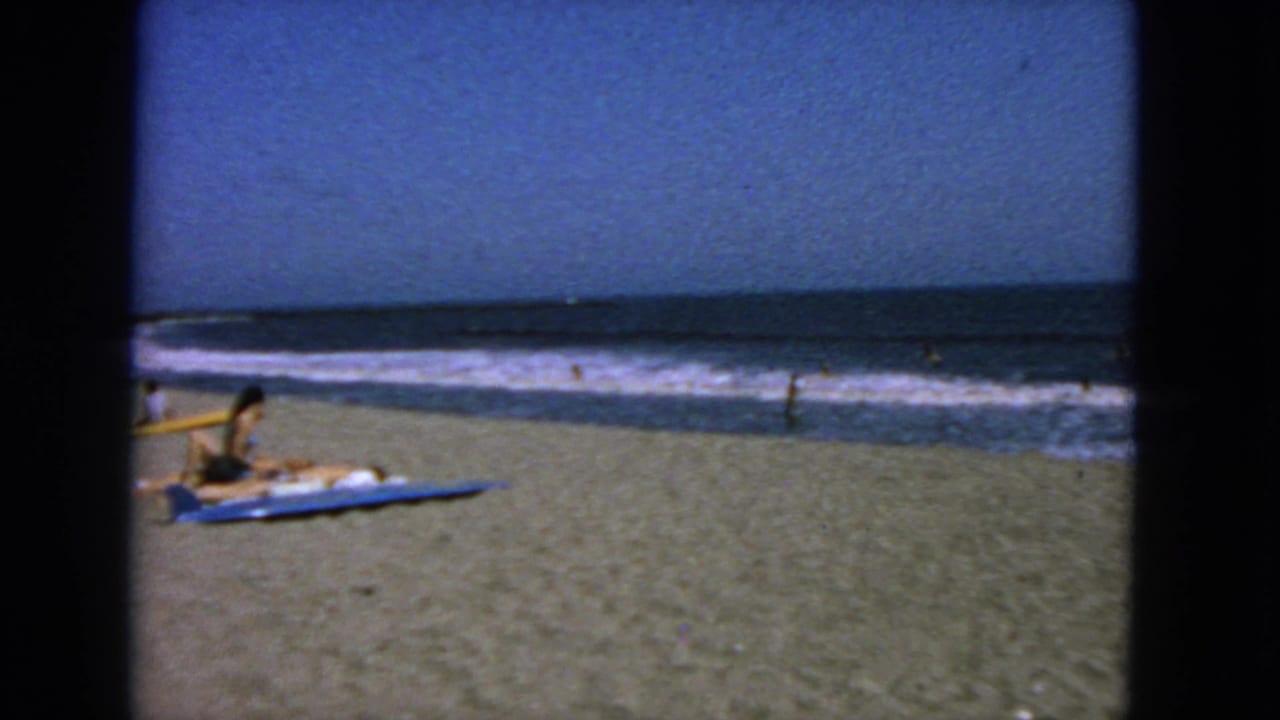 1963 San Francisco beach scene with a person on a blue towel, distant swimmers, surf, and dark window-like bars in the foreground.