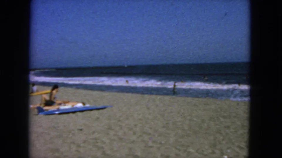 1963 San Francisco beach scene with a person on a blue towel, distant swimmers, surf, and dark window-like bars in the foreground.