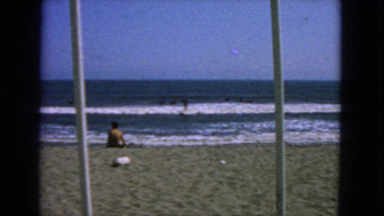 1963 San Francisco shore view with a seated person near the surf, swimmers in the water, and vertical foreground bars crossing the frame.