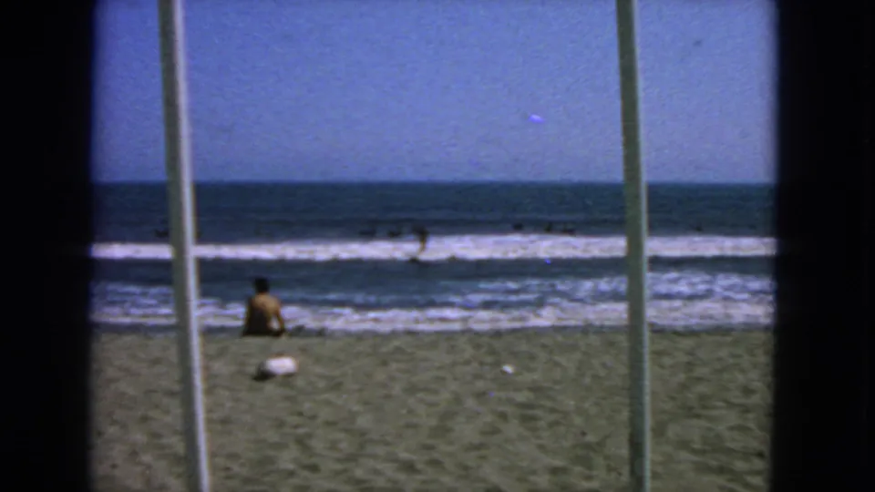 1963 San Francisco shore view with a seated person near the surf, swimmers in the water, and vertical foreground bars crossing the frame.