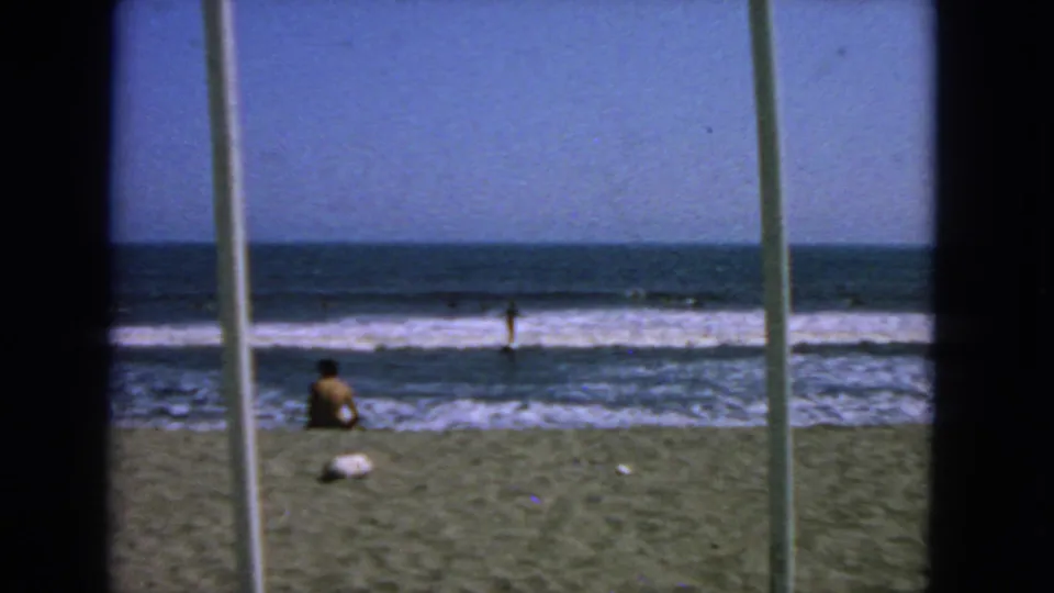 1963 San Francisco beach footage showing a seated person near the waterline, swimmers in the surf, and a dark foreground frame.