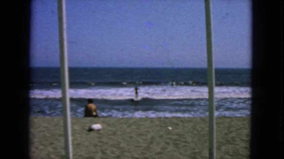 1963 San Francisco shoreline scene with a seated beachgoer, rolling surf, distant swimmers, and window-like vertical foreground framing.
