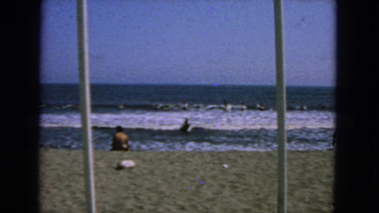 1963 San Francisco beach view through vertical framing, showing a seated beachgoer and distant surfers in low surf.