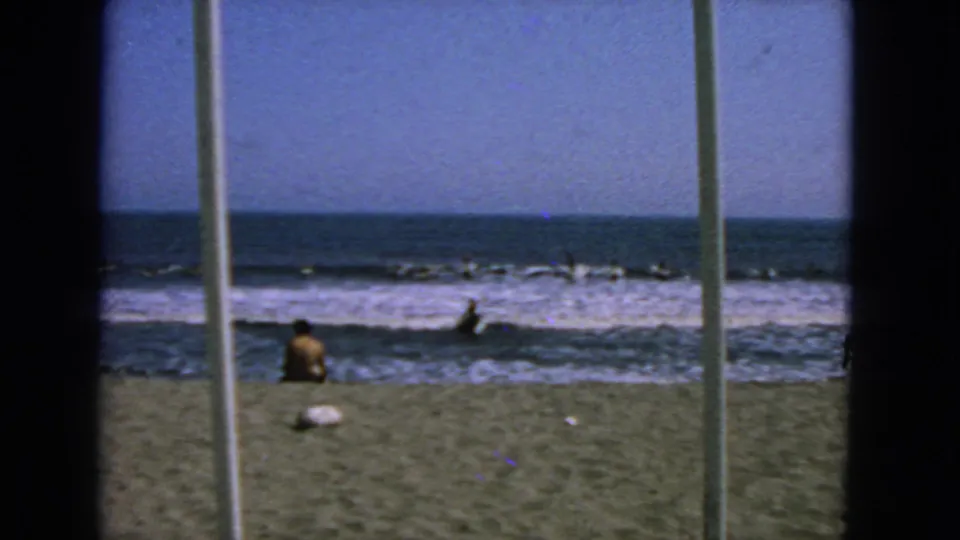 1963 San Francisco beach view through vertical framing, showing a seated beachgoer and distant surfers in low surf.