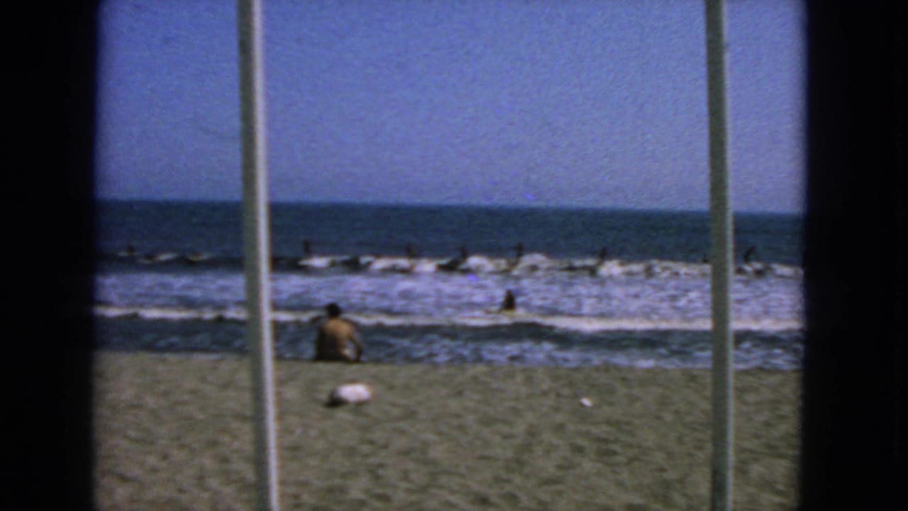 1963 San Francisco surf scene with distant figures in the water and a seated beachgoer near the shoreline.