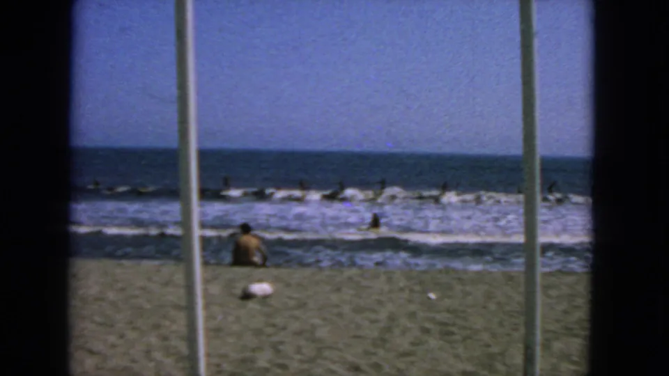 1963 San Francisco surf scene with distant figures in the water and a seated beachgoer near the shoreline.