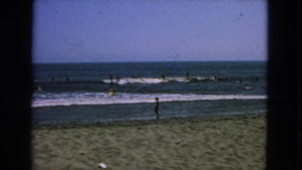 1963 San Francisco beach footage showing a standing person on sand with surfers spread across the small waves.