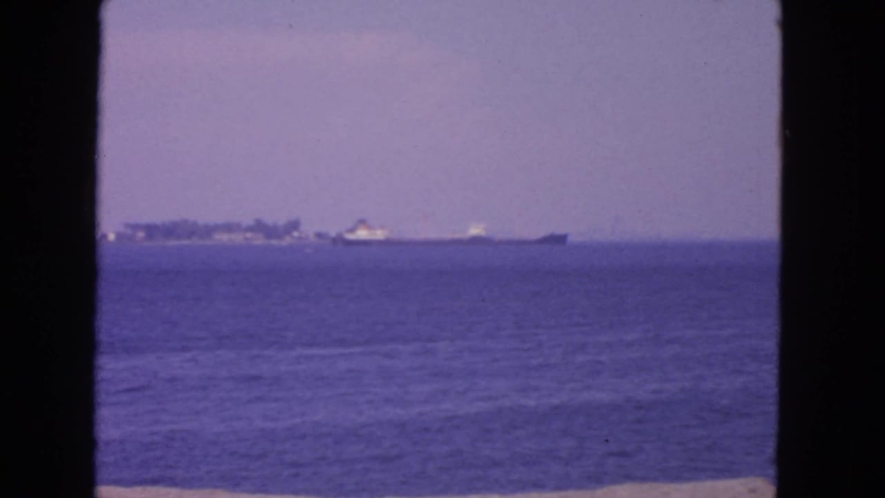 1963 San Francisco Bay home-movie frame showing a distant vessel near a low shoreline across calm blue water.