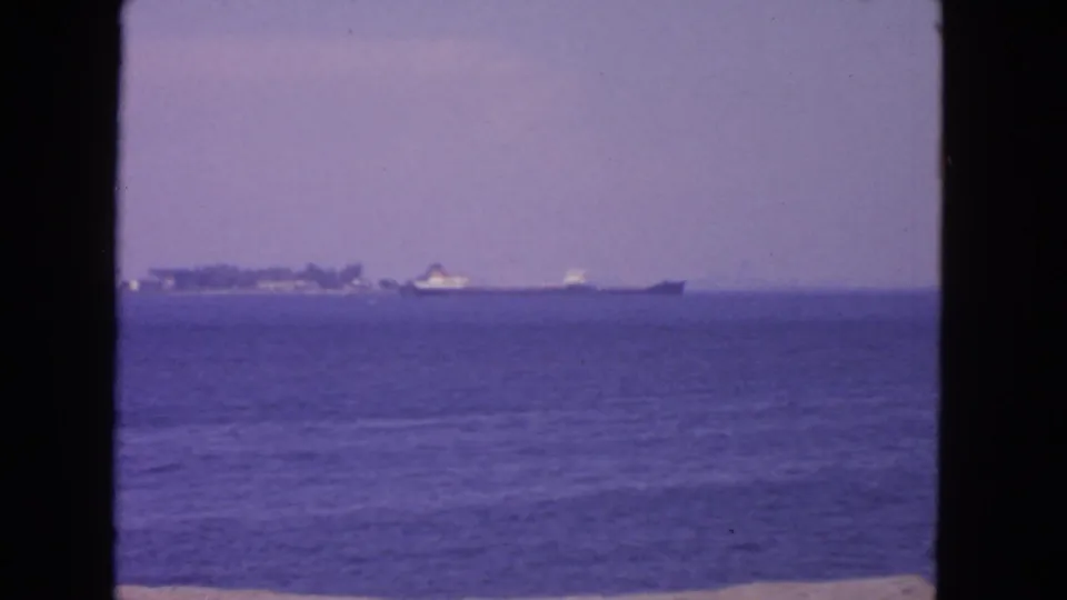 1963 San Francisco Bay home-movie frame showing a distant vessel near a low shoreline across calm blue water.