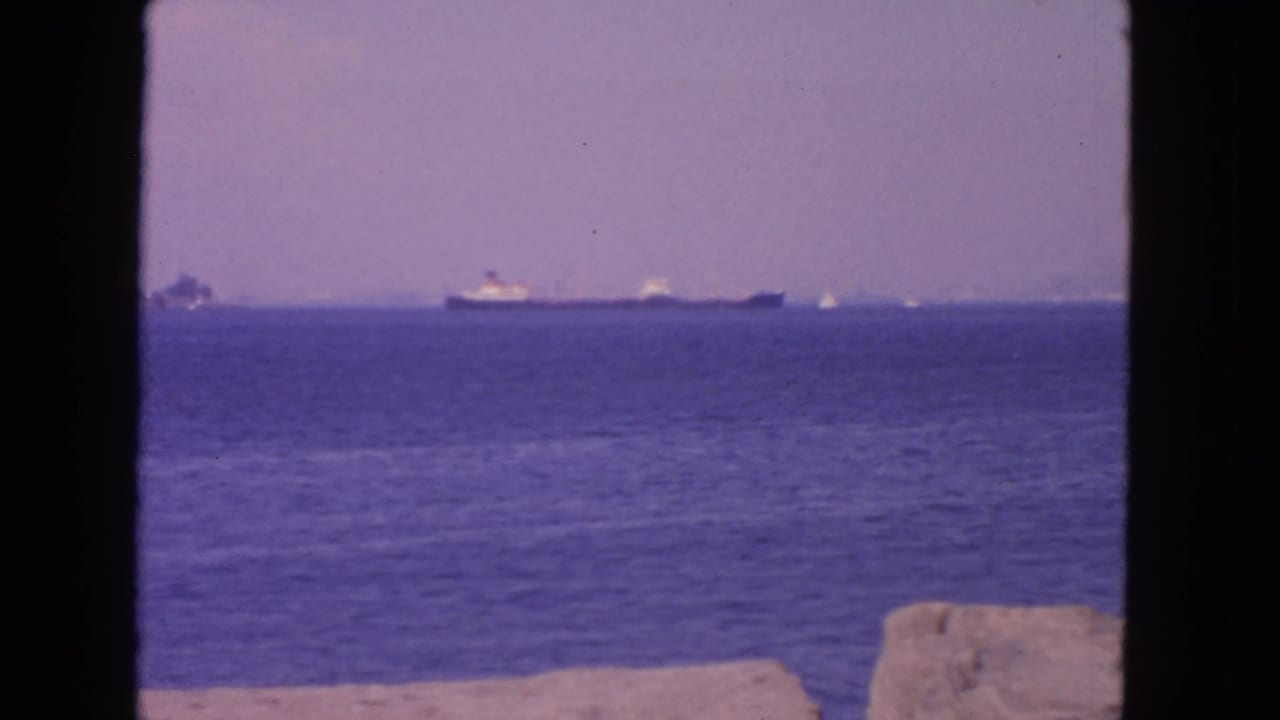 1963 San Francisco bay view with distant cargo ship, small boats, hazy sky, shoreline rocks, and dark foreground framing.