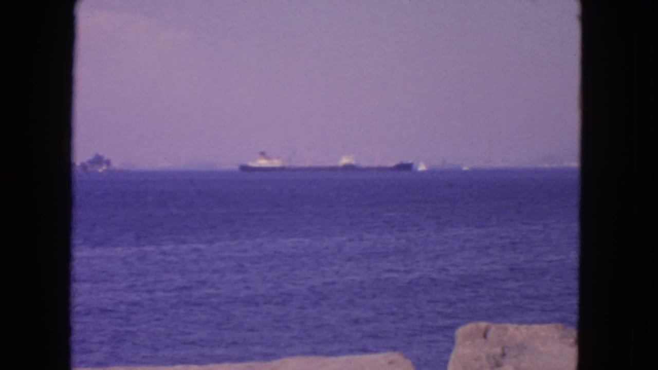 Distant ships cross San Francisco bay water in a 1963 home-movie frame, with soft focus, purple haze, and dark foreground borders.