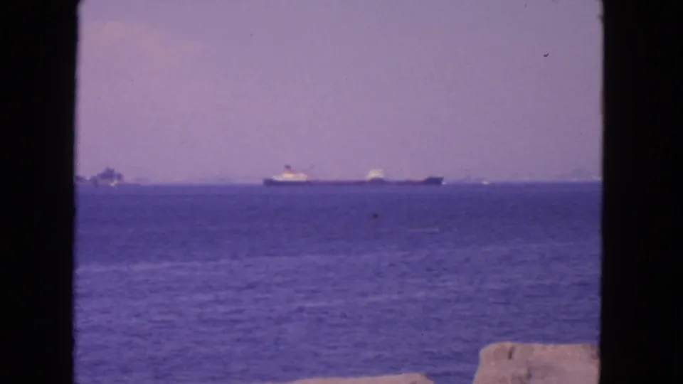 1963 San Francisco bay scene showing a distant cargo ship, smaller vessels, blue water, low rocks, and dark home-movie framing.