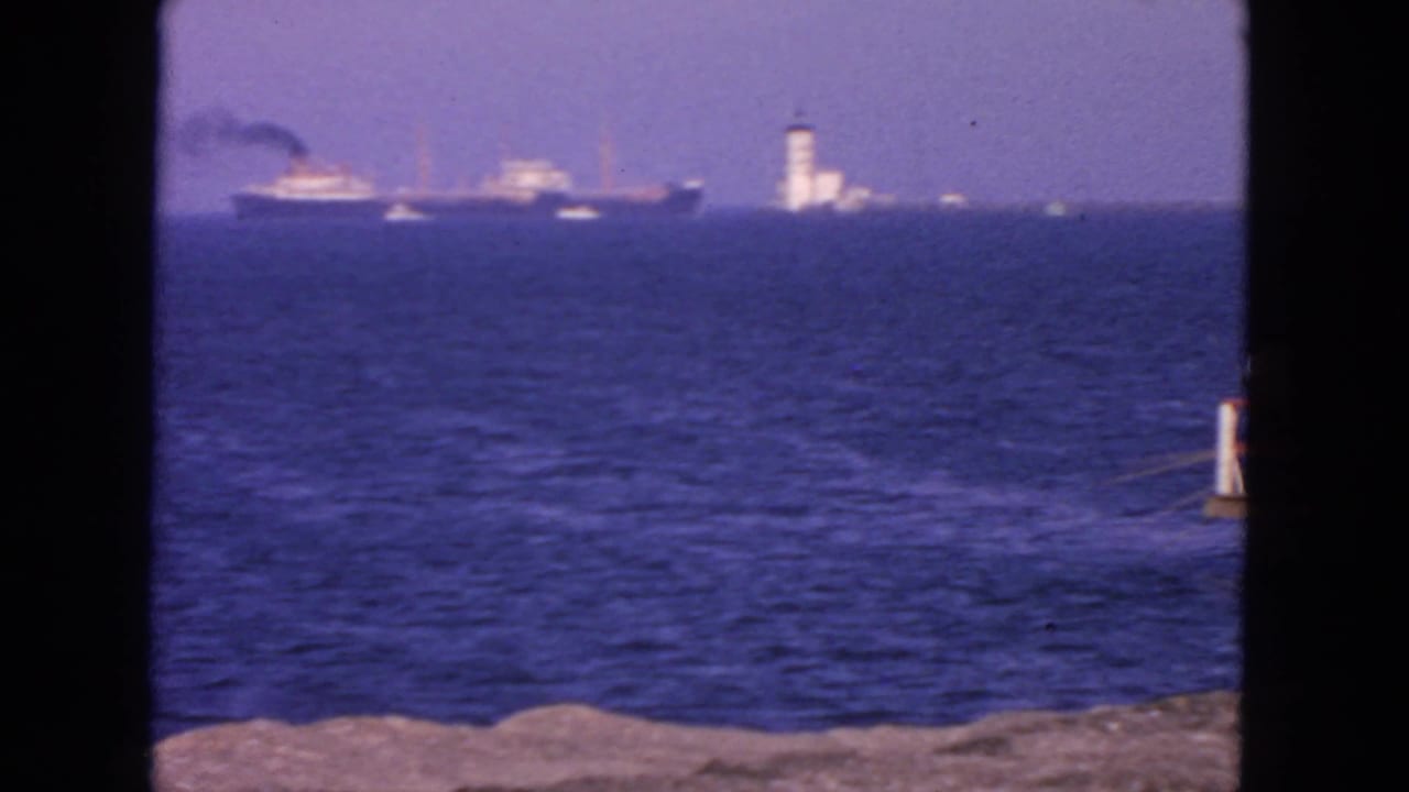 1963 San Francisco Bay view with a distant vessel, a white lighthouse-like tower, blue water, and rocky foreground.