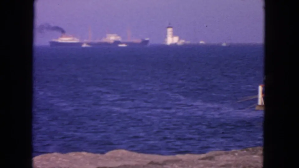1963 San Francisco Bay view with a distant vessel, a white lighthouse-like tower, blue water, and rocky foreground.