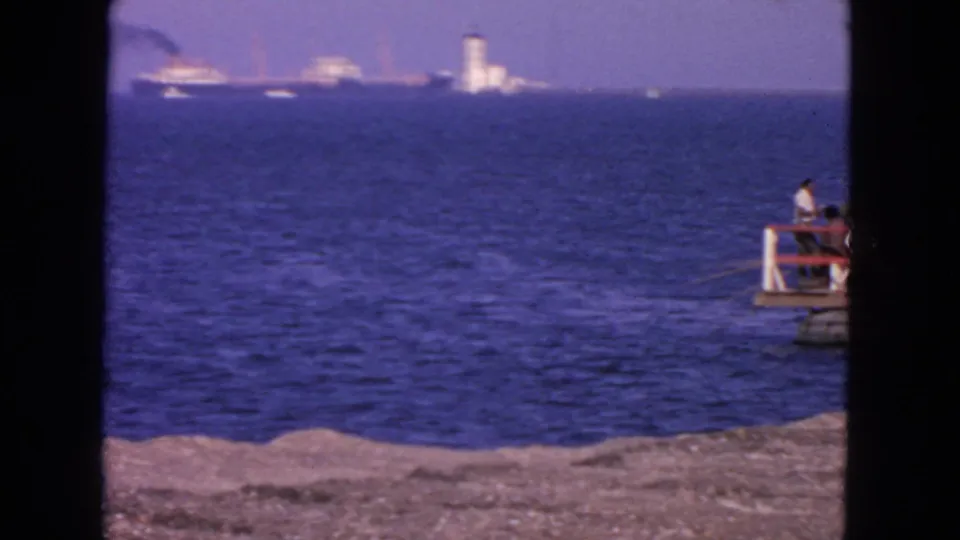 1963 San Francisco Bay home-movie frame with distant shipping, a white tower, rocky shore, and a person at the right edge.